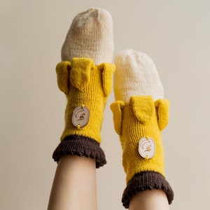 Pode incluir: Um par de meias de tricô amarelo e branco com punhos marrons e etiquetas de madeira. As meias têm um design único com uma ponta pontiaguda e uma pequena aba na parte superior.