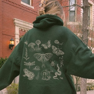 May include: A dark green hooded sweatshirt with a white print of various forest creatures, including mushrooms, butterflies, a dragonfly, a frog, a snail, a beetle, and a snake. The text "Forest" is printed on the bottom right corner of the image.