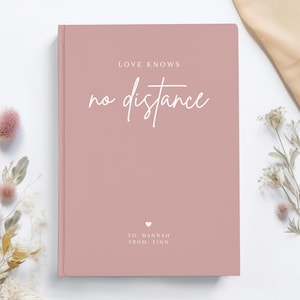 May include: A blush pink hardcover journal with the text "LOVE KNOWS no distance" in white script. The journal has a small heart and the text "TO: HANNAH FROM: FINN" in white.