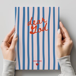 Puede incluir: Un cuaderno rectangular con fondo rosa claro y rayas verticales azules. El texto "dear God" está escrito en cursiva roja en el centro. Debajo, el nombre "HEATHER" está impreso en rojo, seguido de un pequeño icono de un corazón rojo.