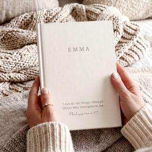 Pode incluir: Um livro de capa dura na cor creme com o nome EMMA impresso em letras maiúsculas cinza-escuras no topo. Abaixo do nome, o texto diz I can do all things through Christ who strengthens me. Philippians 4:13 em uma fonte cinza-escura.