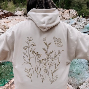 May include: Sand-colored hoodie with a floral and butterfly design on the back. The design features delicate line art of various flowers and a butterfly. The hoodie has a hood and long sleeves. The word "Sand" is printed at the bottom right.