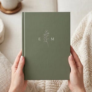 May include: A sage green hardback journal featuring a centred white line art illustration of a delicate floral stem flanked by the capital letters E and M. The notebook has a smooth, matte finish and a clean, rectangular design.