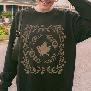 May include: Black sweatshirt featuring a brown bird and floral design within a square frame. The design includes leaves, flowers, and acorns. The word "Black" is printed at the bottom right corner of the sweatshirt.
