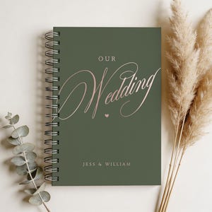 May include: A spiral-bound wedding guest book with a dark green cover. The cover features the words "Our Wedding" in elegant pink script, with the names "Jess & William" at the bottom. Dried ornamental grass and eucalyptus sprigs are in the background.