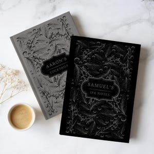 May include: Two hardcover journals with botanical line art patterns. The gray journal features the text "AARON'S COCKTAILS" in a central label, while the black journal features the text "SAMUEL'S IPA NOTES" in a matching central label.