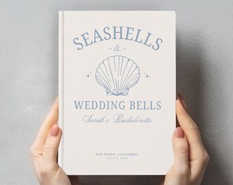 Könnte beinhalten: Ein cremefarbenes Notizbuch mit blauem Text und einer Muschelgrafik. Auf dem Einband steht SEASHELLS & WEDDING BELLS, Sarah's Bachelorette, SAN DIEGO, CALIFORNIA, JULY 6, 2025. Das Design enthält kleine Sternsymbole neben der Muschel.