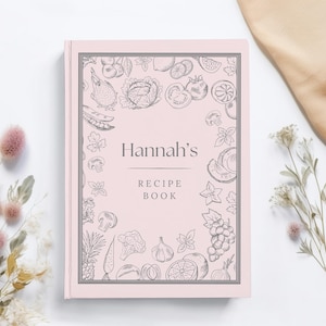 Puede incluir: Un libro de recetas rosa claro con el nombre "Hannah's" en la portada. La portada está decorada con un borde de frutas y verduras esbozadas. Las palabras "Recipe Book" están impresas debajo del nombre. El libro está sobre una superficie blanca.