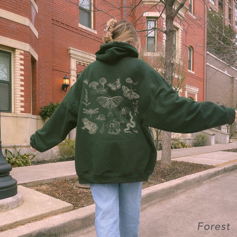May include: A dark green hooded sweatshirt with a white print of various forest creatures, including mushrooms, butterflies, a dragonfly, a frog, a snail, a beetle, and a snake. The text "Forest" is printed on the bottom right corner of the image.