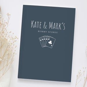 Op de afbeelding: Een marineblauw notitieboek met de tekst "KATE & MARK'S RUMMY SCORES" in witte letters. Een witte illustratie van speelkaarten staat onder de tekst. Het notitieboek ligt op een witte ondergrond met gedroogde bloemen.