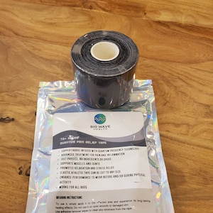 Puede incluir: Un rollo de cinta atlética negra se encuentra sobre una bolsa plateada. La bolsa contiene texto que dice "Quantum Pain Relief Tape" y enumera características como tratamiento avanzado del dolor y soporte muscular. La cinta está diseñada para cortarse a cualquier tamaño.