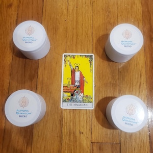 May include: A tarot card reading with four white containers with the text "Aurora Quantum Micro" on them. The tarot card is The Magician, the first card in the Major Arcana. The card is yellow with a red and white illustration of a man holding a wand.