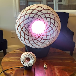 Pode incluir: Duas lâmpadas esféricas geométricas brancas e cobre com uma luz rosa incandescente que emana do centro de cada esfera. A esfera maior está sobre uma base preta e a esfera menor está sobre uma base preta com um cabo.