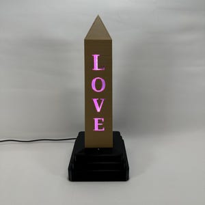 May include: A gold-coloured obelisk with the word "LOVE" cut out and illuminated with pink light. The obelisk is on a black base with three tiers.