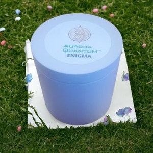 May include: A light blue cylindrical container with a white label that reads "Aurora Quantum™ Enigma". The container is sitting on a white surface with a few purple flowers.