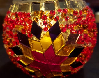 May include: A small, round, glass candle holder with a mosaic design. The mosaic is made of small, colorful glass tiles in shades of red, yellow, gold, and black. The candle holder has a clear glass base and a metal rim.