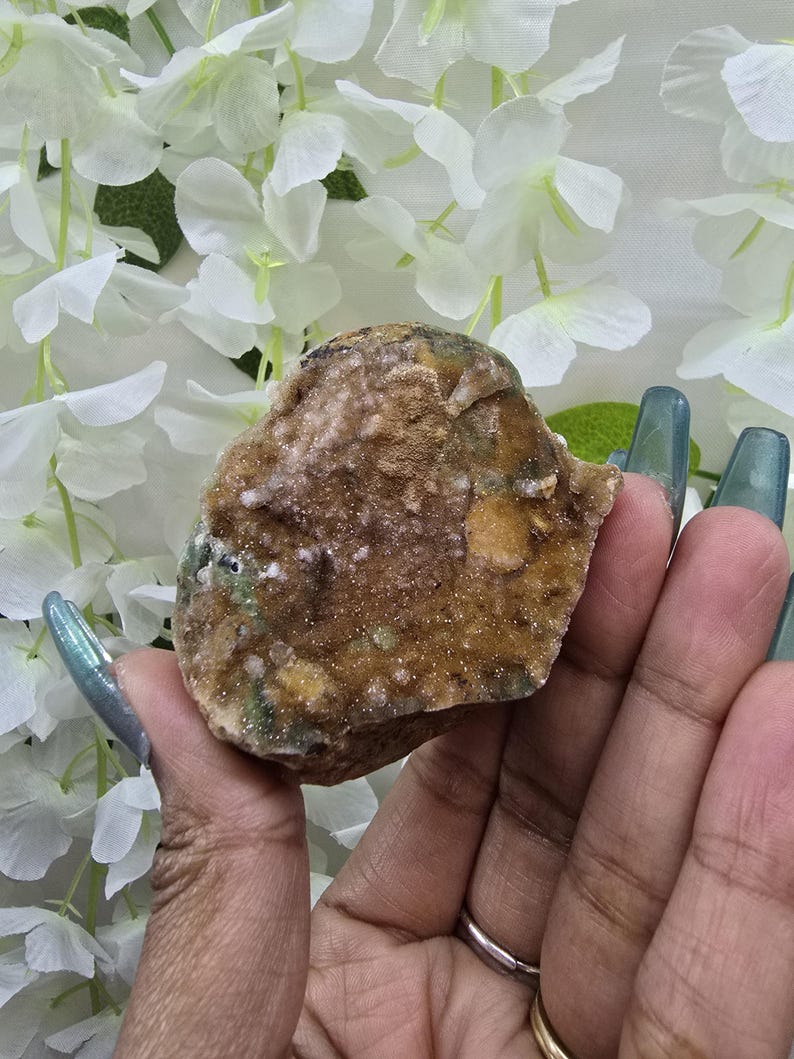May include: A rough, natural crystal formation with a mix of brown, tan, and green hues. The crystal has a textured surface and is held in a hand, with white flowers in the background.