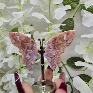 May include: A decorative butterfly figurine with pink and brown patterned wings. The butterfly is mounted on a gold-colored metal stand. The background features white flowers and green leaves.