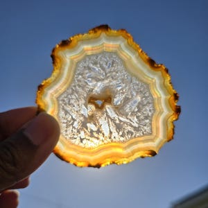 May include: A translucent agate slice with concentric bands of color, including white, yellow, and brown. The natural stone is held up against a blue sky, showcasing its intricate crystalline structure and unique patterns. The agate slice is a decorative item.
