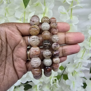 May include: Three beaded bracelets featuring polished, spherical stones in varying shades of brown, tan, beige, and grey. The beads exhibit natural inclusions and marbled patterns, arranged in three distinct strands of different bead sizes.