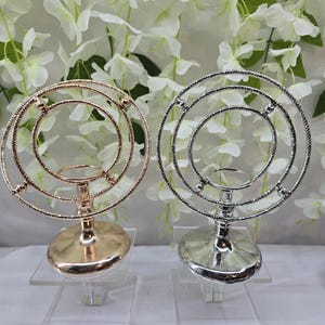 May include: Two decorative metal candle holders. One is gold-toned, the other silver-toned. Each has a circular design with small spheres and a base for a candle. They sit on clear acrylic stands against a backdrop of white flowers.