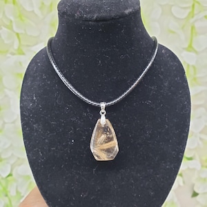 May include: A pendant necklace featuring a teardrop-shaped, translucent gemstone with golden inclusions. The pendant is suspended from a black cord necklace and displayed on a black mannequin. The gemstone is approximately 1.5 inches in length.