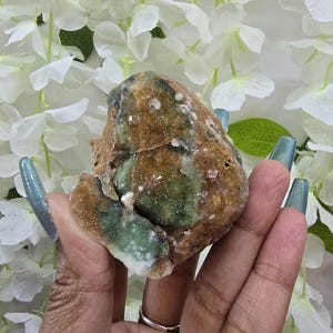 May include: A rough-cut, natural crystal cluster with a mix of brown, green, and white hues. The crystal is held in a hand, showcasing its irregular shape and textured surface. The background features white flowers.