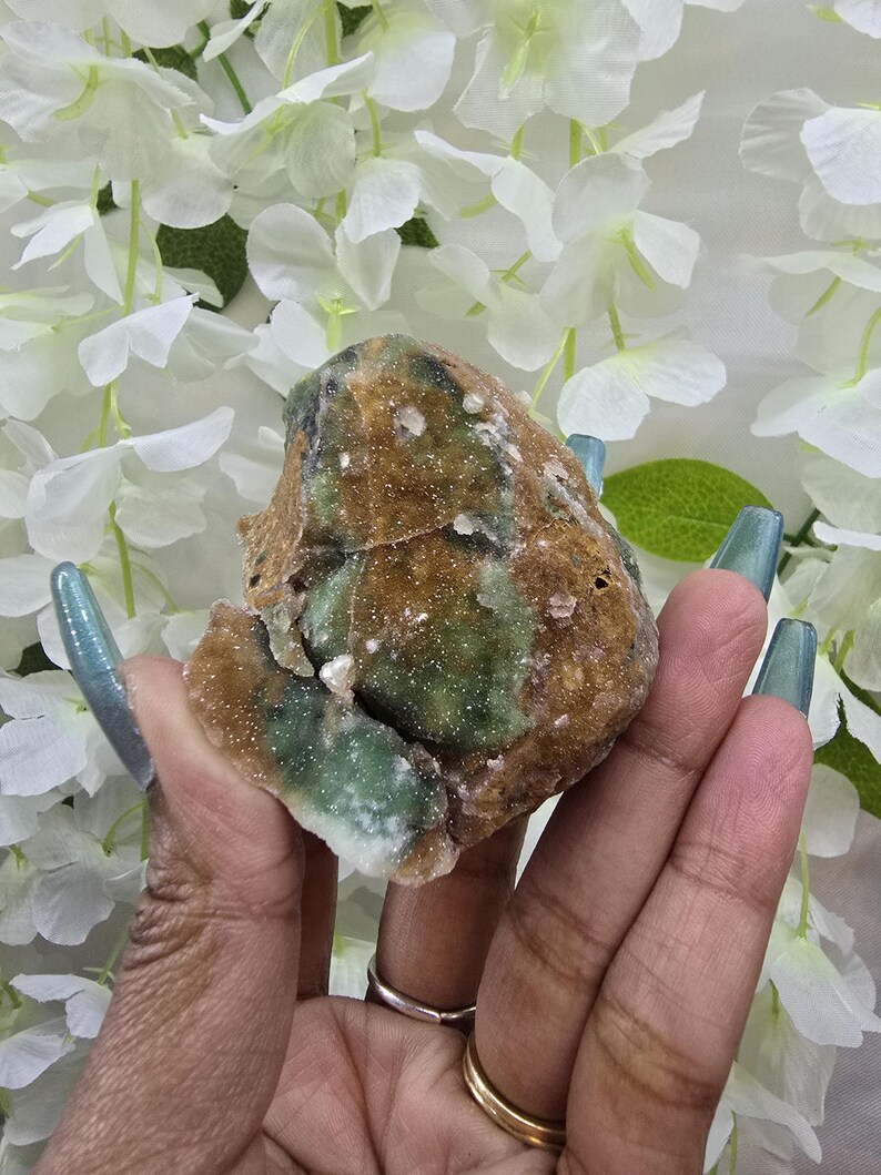 May include: A rough-cut, natural crystal cluster with a mix of brown, green, and white hues. The crystal is held in a hand, showcasing its irregular shape and textured surface. The background features white flowers.