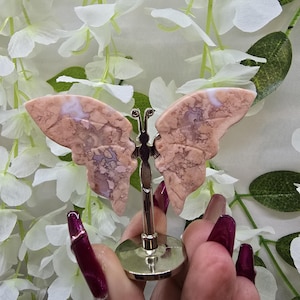 May include: A decorative butterfly figurine with pink and brown patterned wings. The butterfly is mounted on a silver-colored metal stand. The wings have a natural stone appearance. The figurine is displayed against a backdrop of white flowers and green leaves.