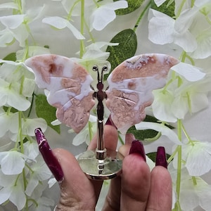 May include: A decorative butterfly figurine with white and pink patterned wings. The butterfly is mounted on a silver-colored metal stand. The wings have a natural stone appearance. The background features white flowers.
