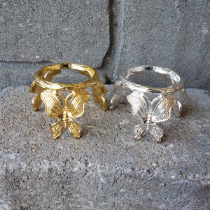 May include: Two decorative metal butterfly stands, one gold and one silver, with a round top and three legs. The stands are designed to hold a crystal ball or other small object.
