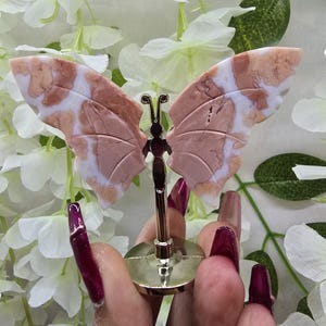May include: A decorative butterfly figurine with pink and white patterned wings, mounted on a gold-colored stand. The butterfly's wings are detailed with natural stone patterns. The figurine is displayed against a backdrop of white flowers and green leaves.