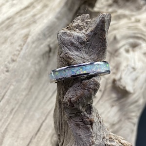 May include: A silver ring with a white opal inlay. The ring is displayed on a piece of driftwood.