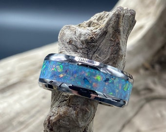 May include: A silver ring with a blue and green opal inlay. The ring is set on a piece of driftwood.