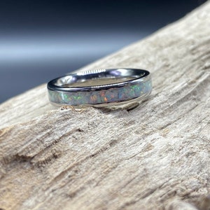 May include: A silver ring with a band of white opal inlay. The ring is on a piece of driftwood.
