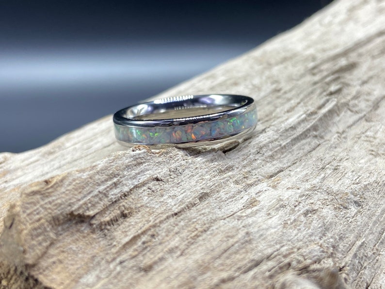 May include: A silver ring with a band of white opal inlay. The ring is on a piece of driftwood.