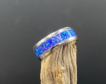 May include: A silver ring with a blue opal inlay. The ring is on a piece of driftwood.
