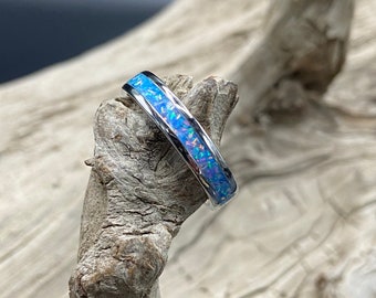 May include: A silver band ring with a blue opal inlay. The ring is displayed on a piece of driftwood.