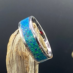 May include: A silver ring with a blue and green opal inlay. The ring is set on a piece of driftwood.