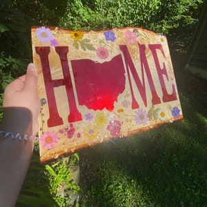 May include: A wooden sign with the word "HOME" spelled out in red letters. The letters are outlined in gold and the background is a light wood with a variety of pressed flowers and gold glitter. The state of Ohio is also outlined in red in the center of the sign.