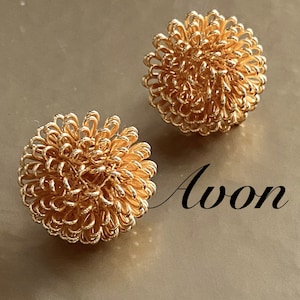 May include: A pair of gold-toned earrings with a flower-like design. The earrings are made of wire that is twisted into a spiral shape. The earrings are labeled "Avon".