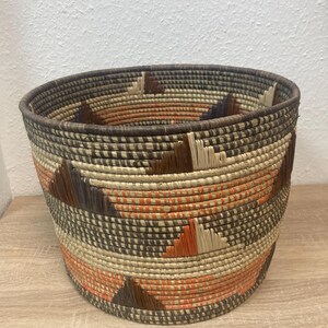 May include: Woven basket with a round shape and a wide opening. The basket features a geometric pattern with triangles in shades of brown, orange, and beige. The rim and base are a darker brown.