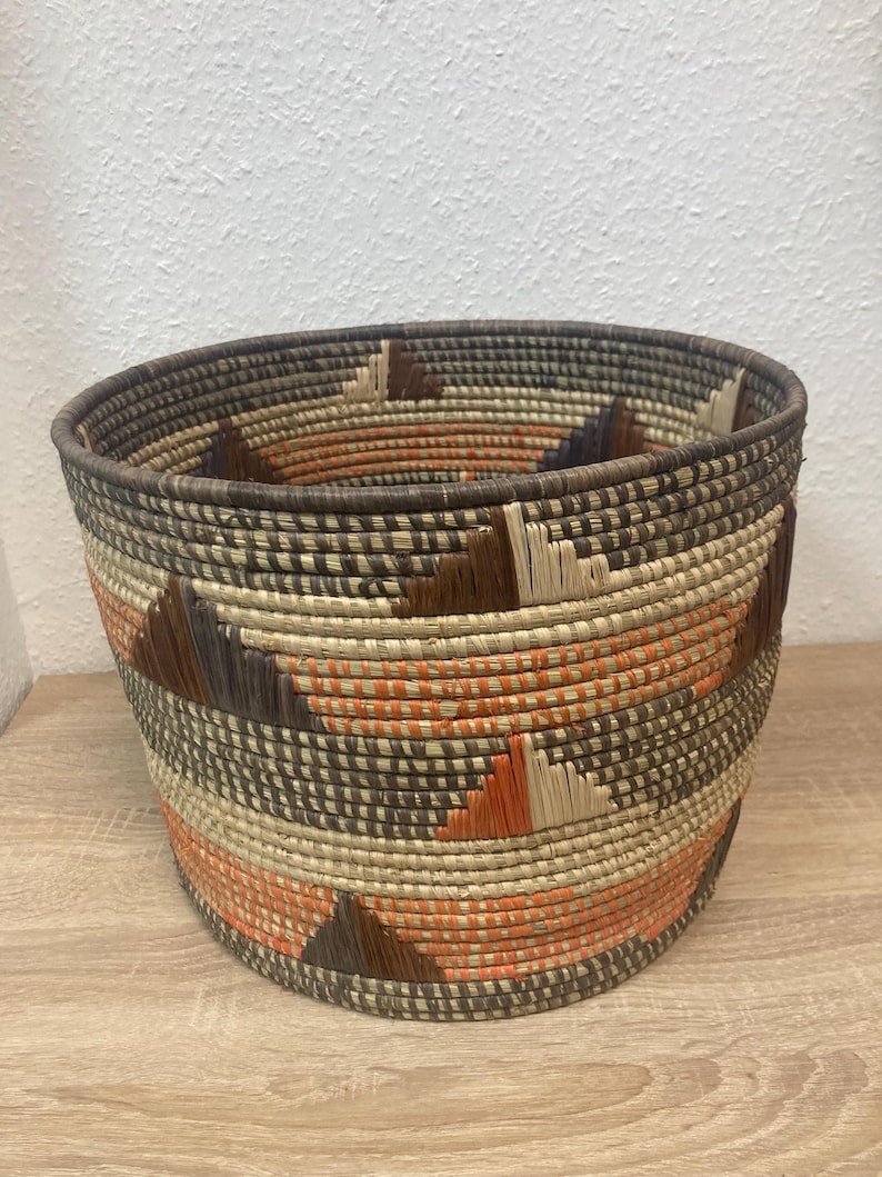 May include: Woven basket with a round shape and a wide opening. The basket features a geometric pattern with triangles in shades of brown, orange, and beige. The rim and base are a darker brown.