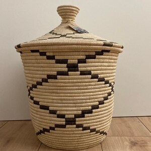May include: A woven basket with a lid, featuring a natural beige color with dark brown geometric patterns. The basket has a cylindrical shape and a matching lid with a small knob. A tag is attached to the lid.