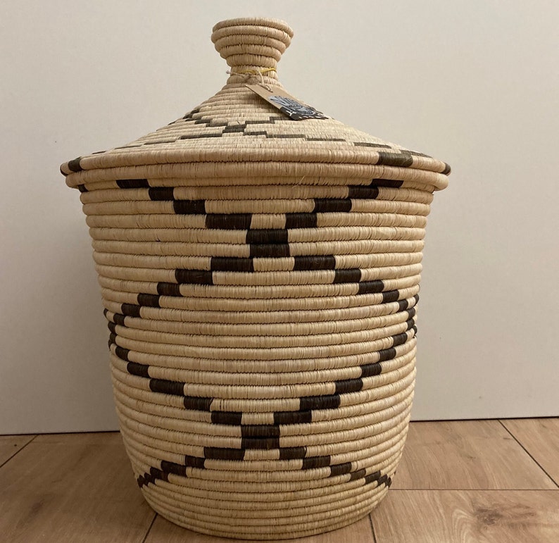 May include: A woven basket with a lid, featuring a natural beige color with dark brown geometric patterns. The basket has a cylindrical shape and a matching lid with a small knob. A tag is attached to the lid.