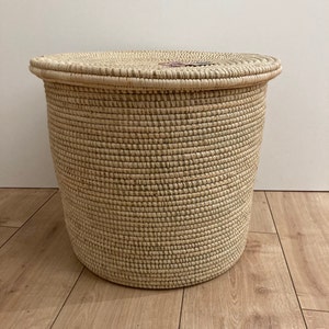 May include: A round woven storage basket with a lid. The basket is made from natural-coloured fibres, with a darker pattern woven throughout. The lid fits securely on top, creating a closed container. The basket is suitable for storage.
