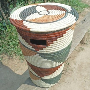 May include: A cylindrical woven basket with a lid. The basket features a color pattern of brown, white, green, and tan. The lid has a circular design. The basket has a rectangular opening on the side.