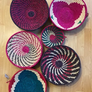 May include: A collection of woven decorative baskets in various sizes and colors. The baskets feature spiral and geometric patterns in shades of pink, teal, green, and white. The baskets are made of natural materials and have a rustic, handmade appearance.