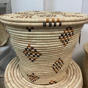 May include: A woven basket with a lid. The basket is cylindrical with a slightly flared top. It is made of natural-colored fibers with geometric patterns in brown and black. The basket is sitting on a smaller, round woven mat.