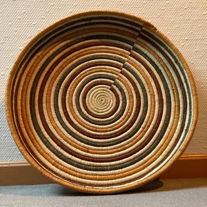 May include: A round woven basket with a spiral design. The basket features concentric circles of varying colors, including beige, brown, orange, and dark green. The rim and outer edge are a darker brown. The basket is likely used for decorative or storage purposes.